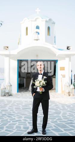 Stilvoller Bräutigam wartet vor der Kirche auf die Braut mit einem Blumenstrauß Stockfoto