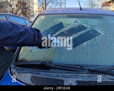 Autofenster im Winter reinigen. Der Mensch kratzt mit einem Plastikkratzer von der Windschutzscheibe eines blauen Autos. Nahaufnahme. Selektiver Fokus. Stockfoto