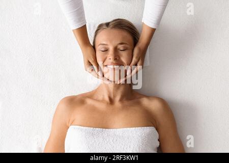 Therapeut macht Lifting-Gesichtsmassage für wunderschöne Dame mittleren Alters, Draufsicht Stockfoto
