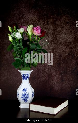 Frische Schnittblumen in einer weißen und blauen Keramikblütenvase mit Buch auf einem Holztisch Stockfoto