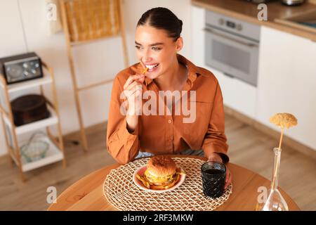 Fröhliche junge Frau, die pommes frites und Hamburger hält und isst, am Tisch im Küchenbereich sitzt, freier Platz Stockfoto