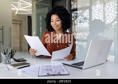Junger erfolgreicher Finanzier arbeitet mit Dokumenten und Verträgen im Büro, zufrieden mit den Ergebnissen der Finanzaktivitäten, Geschäftsfrau prüft Konten, Berichte und Verträge, Laptop. Stockfoto