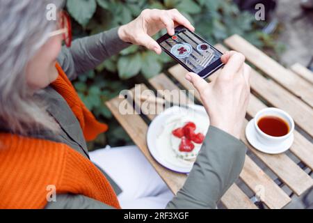 Eine ältere Dame fotografiert das Erdbeerdessert mit dem Smartphone am Tisch im Freien Stockfoto