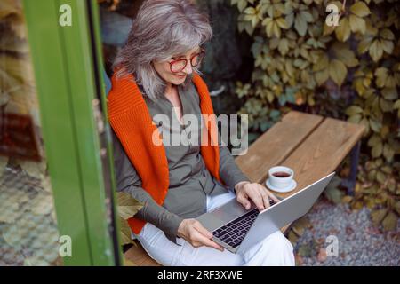 Lächelnde reife Frau arbeitet auf einem Laptop, der auf einer Bank neben dem Cafefenster sitzt Stockfoto