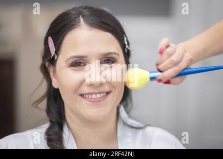 Nahaufnahme der Make-up-Bürste, die Puder auf die lächelnde Braut aufträgt, um am Morgen ihrer Hochzeit zu sein Stockfoto