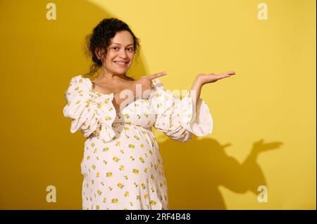 Fröhliche, lockige, multiethnische, schwangere Frau in stilvollem Sommerkleid, zeigt mit dem Zeigefinger auf die Werbetafel auf ihrer Hand, lächelt Stockfoto