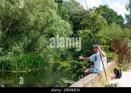 Angler sitzt neben dem River Stour in Flatford in der Nähe von John Constable, der an einem heißen Sommertag 2023 den Hay Wain malte - Suffolk, England, Großbritannien Stockfoto