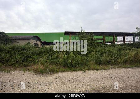 Lagerhaus hinter einem Zaun und Büschen an einem bewölkten Tag in der italienischen Landschaft, von einer unbefestigten Straße aus gesehen Stockfoto