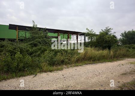 Lagerhaus hinter einem Zaun und Büschen an einem bewölkten Tag in der italienischen Landschaft, von einer unbefestigten Straße aus gesehen Stockfoto