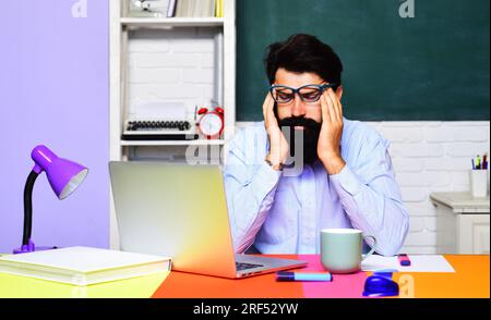 Erschöpfter gestresster Lehrer mit Kopfschmerzen am Schreibtisch im Klassenzimmer. Müde männliche Schülerin, die sich nach einer riesigen Menge Computer Migräne fühlt Stockfoto