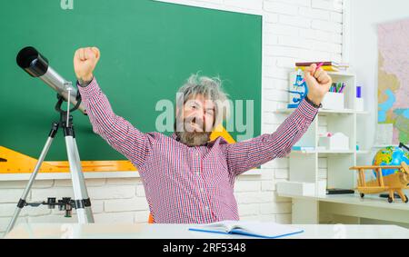 Aufgeregter Lehrer am Arbeitsplatz. Lehrertag. Glücklicher, bärtiger Professor am Tisch im Klassenzimmer. Lernen und Bildung. Zurück in die Schule. Schüler in Stockfoto