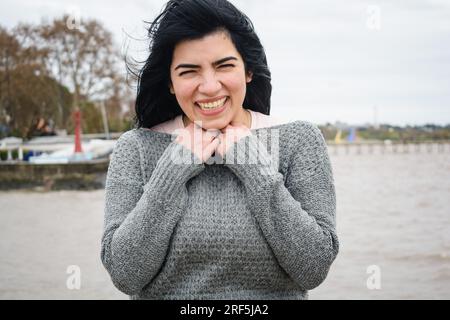 Taille hoch Porträt einer jungen lateinischen Touristin venezolanischer Herkunft in einem grauen Pullover, sie ist kalt und steht glücklich auf dem Pier und schaut sich an Stockfoto