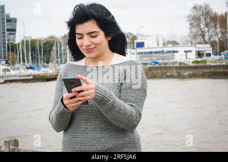 Porträt einer jungen lateinischen Touristin venezolanischer Herkunft, lächelnd auf dem Pier mit dem Fluss im Hintergrund, mit ihrem Handy, sendend Stockfoto