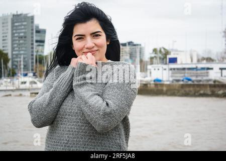 Taille hoch Porträt einer jungen lateinischen Touristin venezolanischer Herkunft in einem grauen Pullover, sie ist kalt und steht glücklich auf dem Pier und schaut sich an Stockfoto