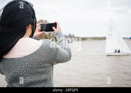 Hüfthoher Rückblick auf die junge, nicht wiedererkennbare Touristin, die mit ihrem Handy auf dem Pier steht und ein Foto von den Booten auf dem Fluss macht, Technologie und Stockfoto