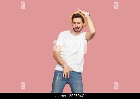 Porträt eines aufgeregten, verspielten, gutaussehenden Mannes mit weißem T-Shirt und Hut, der lustige Posen hat und Zeit auf einer Party verbringt. Studio-Aufnahme im Innenbereich isoliert auf pinkfarbenem Hintergrund. Stockfoto