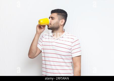 Porträt eines gutaussehenden jungen bärtigen Mannes, der gestreiftes T-Shirt trägt und dabei eine gelbe Tasse hält, Kaffee, Tee oder Wasser trinkt. Studioaufnahmen im Innenbereich isoliert auf grauem Hintergrund. Stockfoto