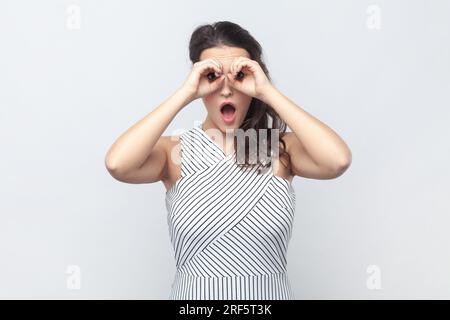 Portrait einer überraschten, interessierten brünetten Frau, die Ferngläser mit den Händen macht und in einem gestreiften Kleid vor der Kamera erstaunt aussieht. Studioaufnahmen im Innenbereich isoliert auf grauem Hintergrund. Stockfoto