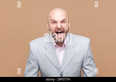 Porträt eines wütenden oder schockierten bärtigen Mannes, der steht, in die Kamera schaut und schreit, sich mit einem Freund streitet, eine graue Jacke trägt. Studioaufnahmen isoliert auf braunem Hintergrund. Stockfoto