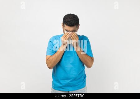 Porträt eines gestressten, unrasierten Mannes, der ein blaues T-Shirt trägt, mit der Hand nach unten steht und weint, Trauer und Traurigkeit ausdrückt. Studioaufnahmen im Innenbereich isoliert auf grauem Hintergrund. Stockfoto