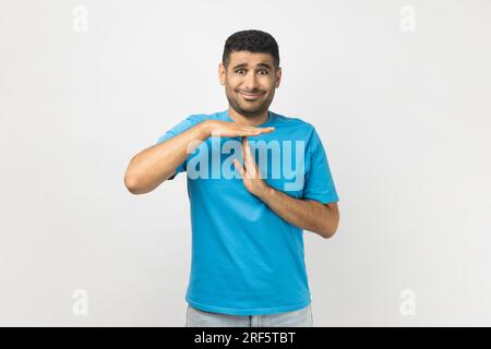 Porträt eines attraktiven jungen, unrasierten Mannes, der ein blaues T-Shirt im Stehen trägt und Auszeit-Geste mit den Händen zeigt, in die Kamera schaut. Studioaufnahmen im Innenbereich isoliert auf grauem Hintergrund. Stockfoto