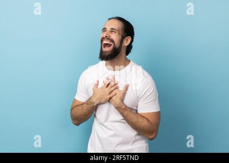 Das Porträt eines fröhlichen Mannes mit Bart und weißem T-Shirt hält die Hände auf der Brust, schließt die Augen und drückt positive Emotionen aus. Innenstudio, isoliert auf blauem Hintergrund. Stockfoto