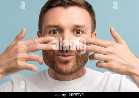 Ein junger Mann, der Spaß hat, Gesichtscreme auf die Wangen zu tragen, zwei Hände, die die Kamera anschauen, und die Nahaufnahme lächelt Stockfoto