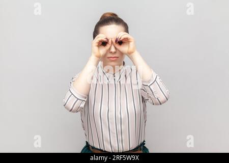 Porträt einer lustigen, positiven, jungen erwachsenen Frau, die ein gestreiftes Hemd trägt, mit binokularen Gesten steht und sich etwas Interessantes ansieht. Studioaufnahmen im Innenbereich isoliert auf grauem Hintergrund. Stockfoto