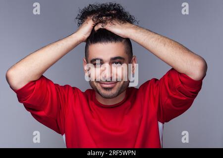 Porträt eines attraktiven, lächelnden, bärtigen Mannes mit lockigem Haar, der roten Pullover trägt, in die Kamera schaut, sein Haar sammelte. Studioaufnahmen im Innenbereich isoliert auf grauem Hintergrund. Stockfoto