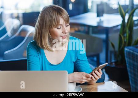 Porträt einer schönen, fröhlichen, optimistischen jungen Frau mit blondem Haar im blauen Hemd, die am Laptop arbeitet, Pause hat, Handy benutzt, soziale Netzwerke überprüft. Indoor-Shot im Café. Stockfoto