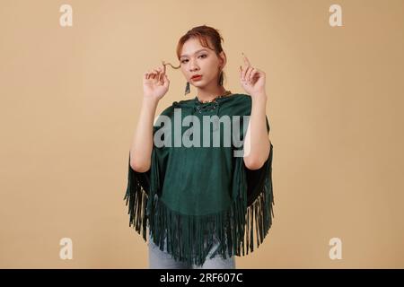 Porträt einer jungen Transgender-Frau im grünen Poncho, die ihr Haar berührt und in die Kamera schaut Stockfoto