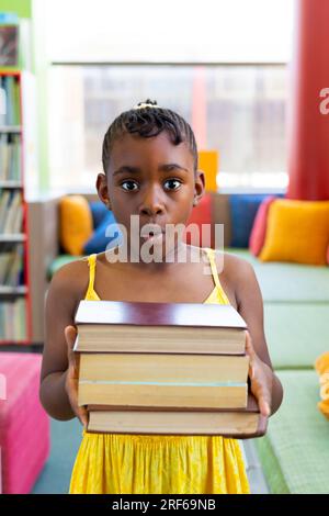 Porträt eines schockierten afroamerikanischen Schulmädchens, das Bücher mit bunten Kissen auf der Couch hält Stockfoto