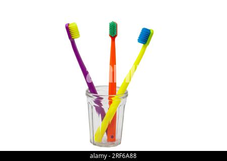 Drei Zahnbürsten – violett, orange und gelb mit verschiedenfarbigen Borsten in einem durchsichtigen Becher auf weißem Hintergrund. Isoliert. Vertikal. Stockfoto
