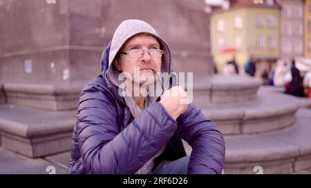 Warschau, Polen. 11. Juni 2023. Porträt eines erwachsenen Mannes mit Brille, der in der Kapuze auf dem Platz sitzt und eine Tabakpfeife auf dem Palastplatz raucht, Warschauer Altstadt (Kreditbild: © Andrey Nekrasov/ZUMA Press Wire), NUR REDAKTIONELLE VERWENDUNG! Nicht für den kommerziellen GEBRAUCH! Stockfoto