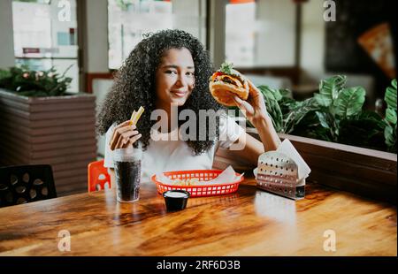 Glückliches Mädchen, das einen köstlichen Hamburger in einem Restaurant genießt. Porträt einer jungen Afro-Frau, die einen Hamburger mit Pommes frites in einem Restaurant hält Stockfoto