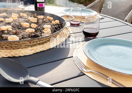 Schwarzer Reis mit Tintenfisch und Garnelen in einer Paella-Pfanne. Serviert mit Rotwein auf der Terrasse Stockfoto