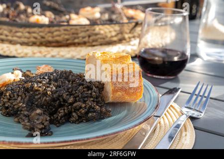 Schwarzer Reis mit Tintenfisch und Garnelen in einer Paella-Pfanne. Fertig zum Essen, mit Brot und Rotwein Stockfoto