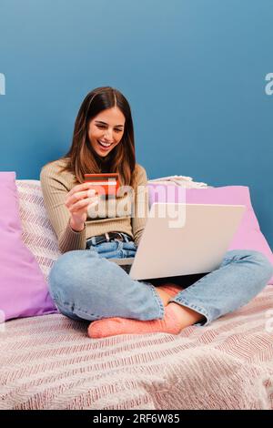 Vertikales Porträt einer jungen Frau, die eine Kreditkarte und einen Laptop benutzt, um einen Lieferauftrag auf der Heimcouch zu bezahlen. Glückliche Frau, die im Internet mit einem Notebook auf dem Sofa einkauft, kauft und einkauft. Hochwertiges Foto Stockfoto