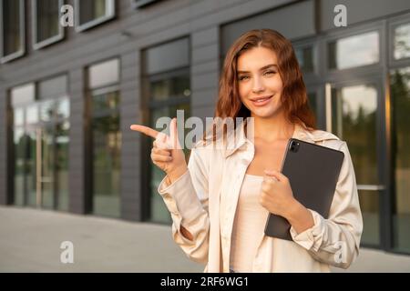 Lächelnde, rothaarige Geschäftsfrau, die ein Tablet hielt und nach links zeigte, in der Nähe des Business Centers. Stockfoto