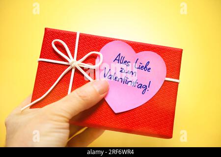 FOTOMONTAGE, Hand hält Geschenk mit einem herzförmigen Zettel und der Aufschrift alles Liebe zum Valentinstag Stockfoto