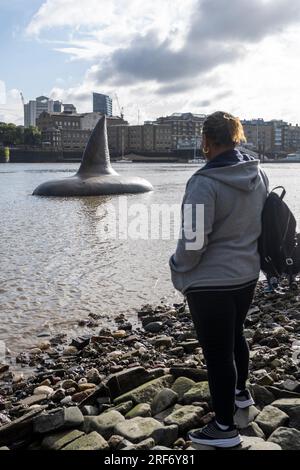 London, Großbritannien. 1. August 2023. Eine Frau beobachtet eine der drei riesigen Megalodon-Haifischflossen, die sich scheinbar an der Oberfläche der Themse in der Nähe der Tower Bridge erheben, um für die Veröffentlichung der Warner Bros. In Großbritannien am 4. August zu werben Filmfilm „Meg 2: The Trench“ mit Jason Statham. Laut dem Naturkundemuseum war die prähistorische Art, bekannt als Otodus megalodon, der größte Hai der Welt und einer der größten Fische, die es je gab. Kredit: Stephen Chung / Alamy Live News Stockfoto