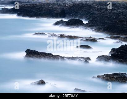 Lange Exposition einer felsigen Küstenlinie. Das Meer hat eine leichte metallische Farbe mit nebligen weißen Rändern an den schwarzen Felsen. Stockfoto