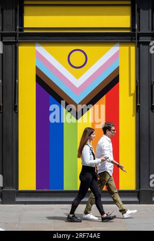 Am 9. Juli 2023 in London, Großbritannien, passieren die Menschen eine „Pride Progress“-Flagge in einem leeren Schaufenster. Die Flagge enthält die Streifen der Regenbogenflagge, die LGBTQ+-Gemeinschaften repräsentieren, mit Farben von der Transgender Pride Flag und auch farbigen Menschen. Stockfoto