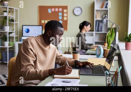 Seitliches Porträt eines schwarzen jungen Mannes, der am Schreibtisch im Büro arbeitet und während der Projektplanung Notizen in Notizbuch schreibt Stockfoto