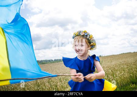 Die ukrainische Flagge ist auf den Schultern eines kleinen ukrainischen Mädchens. Glückliches ukrainisches Kind in freier Ukraine ohne Krieg läuft auf einem Feld entlang einer Landstraße Stockfoto