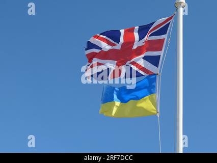 Schließung der Nationalflaggen der Ukraine und des Vereinigten Königreichs, die unter windigen Bedingungen auf einer weißen Fahnenstange vor einem blauen Himmel zu sehen sind. Stockfoto