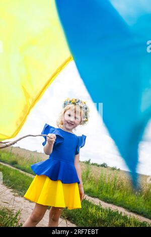 Die ukrainische Flagge ist auf den Schultern eines kleinen ukrainischen Mädchens. Glückliches ukrainisches Kind in freier Ukraine ohne Krieg läuft auf einem Feld entlang einer Landstraße Stockfoto