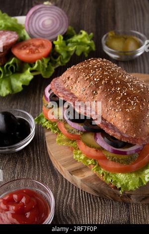 Foto-Sandwich mit frischem Essen auf einem Holzbrett. Wurst, Zwiebeln, schwarze Oliven, frische Salatblätter, Brot auf dem Tisch Stockfoto