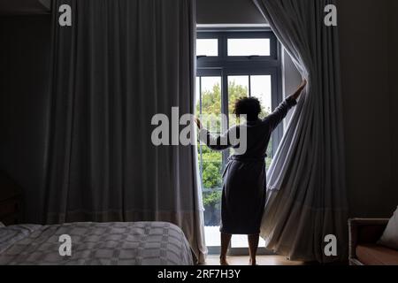 Rücken einer birassischen Frau im Bademantel, die Vorhänge im Schlafzimmer öffnet, Kopierraum Stockfoto