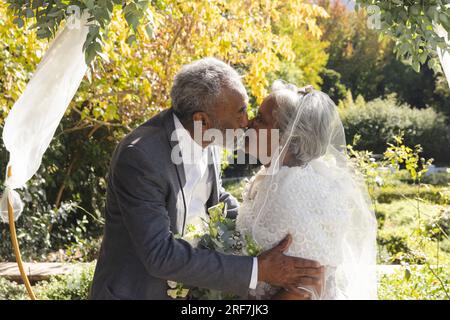 Glückliche ältere, birassische Braut und Bräutigam, die sich bei der sonnigen Hochzeitszeremonie im Freien küssen Stockfoto